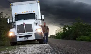 Conductor de camión, con sus obligaciones, posa junto a su vehículo bajo un cielo tormentoso.
