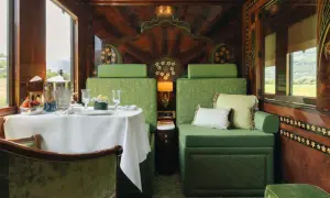 Luxuriöser Speisewagen im Orient Express: Ein gedeckter Tisch mit Obst und Champagner, grüne Sitzmöbel und kunstvolle Holzverzierungen.