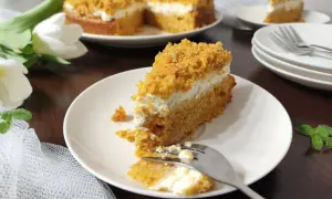 Pyszne ciasto marchewkowe z mascarpone, posypane kruszonką. Na talerzu kawałek z widelcem, obok cały tort i białe tulipany.