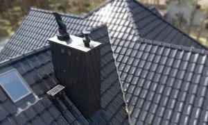 Czarny, nowoczesny komin na dachu z dachówki ceramicznej. Obok okno dachowe i stopień kominiarski.