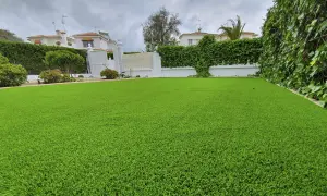 Un jardín con césped artificial impecable, delimitado por vegetación exuberante y casas blancas al fondo.