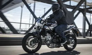 Motocyklista w kasku jedzie srebrnym cafe racerem Suzuki SV650X po moście.