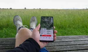 Person sitzt auf einer Holzbank, hält ein Smartphone mit einer guten Wander-App, die eine 6,74 km lange Tour mit 16 hm Anstieg und 672 hm Seehöhe anzeigt.
