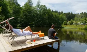 Ein Mann angelt, während eine Frau auf einer Liege entspannt. Perfekter Urlaub in Deutschland am See mit Blick auf den Wald.