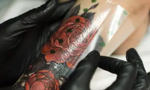Ręce w czarnych rękawiczkach zdejmują folię ochronną z nowego, czerwonego tatuażu z różami. To ważny krok, jak dbać o tatuaż.