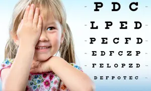 Uśmiechnięta dziewczynka zasłania oko podczas badania wzroku u dzieci. Obok tablica Snellena z literami.