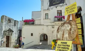 Un vicolo pittoresco a San Michele sul Gargano, con un grande pane rustico in primo piano e cartelli che pubblicizzano "Pane cotto nel forno a legna" e "Panini alla piastra".