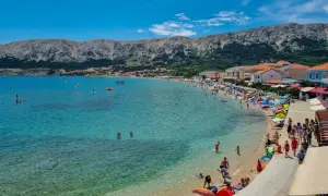Słoneczny dzień na plaży w Baška, Krk, Chorwacja. Turkusowa woda, tłumy ludzi, góry w tle.
