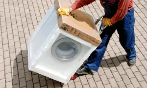 Pracownik w kombinezonie i rękawicach transportuje zapakowaną pralkę na wózku. Na kartonie widnieje napis "MAX 600 kg", co sugeruje, ile waży pralka.