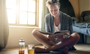 Hombre joven leyendo un libro, disfrutando del **slow living** junto a una ventana con luz natural y una taza de té.