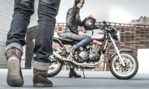 Kobieta w skórzanej kurtce siedzi na motocyklu, obok stoją nogi w brązowych butach motocyklowych.