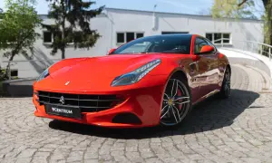 Czerwone Ferrari FF na brukowanej drodze, z budynkiem i drzewami w tle.
