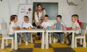 Nauczycielka bez wykształcenia pedagogicznego prowadzi zajęcia z dziećmi w przedszkolu. Dzieci siedzą przy stolikach, bawiąc się klockami i kartami.