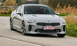Szary Kia Stinger z kierowcą za kierownicą jedzie po krętej drodze, otoczony jesienną roślinnością.