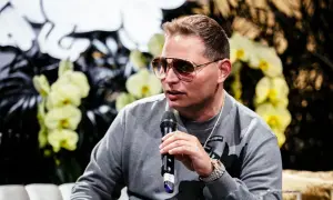 Scott Storch, known for his hit songs, speaks into a microphone, wearing sunglasses and a grey sweatshirt.