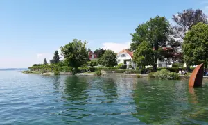 Friedrichshafen am Bodensee: sonnige Uferpromenade mit weißen Villen, grünen Bäumen und einer rostigen Skulptur im Wasser.