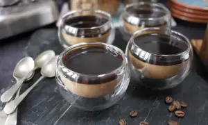 Fyra små glas med kaffe pannacotta, toppad med mörk sirap. Tre skedar ligger till vänster, och kaffebönor sprider sig på stenen.