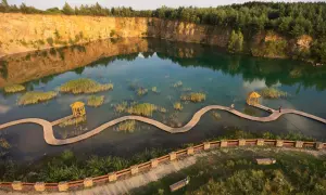 Malowniczy park Gródek z drewnianą kładką wijącą się pośród turkusowej wody i trzcin. W oddali skalne zbocza i las.
