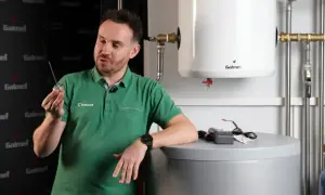Mężczyzna w zielonej koszulce prezentuje element, który może być anodą tytanową, obok zasobnika wody.