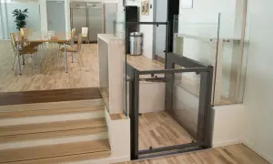 Piattaforma elevatrice per interni con cabina in vetro e struttura metallica, integrata in un ambiente moderno con scale in legno e sala da pranzo.