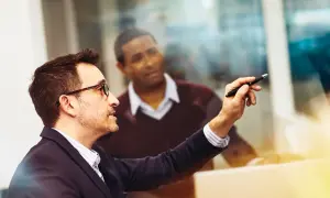 Uomo con occhiali indica un punto su uno schermo, discutendo un **salary review** con un collega in background.