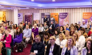 Sala konferencyjna pełna ludzi siedzących w rzędach, z banerami sponsorów w tle.
