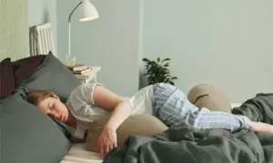 Kvinna sover i sängen, omgiven av kuddar. Hon har på sig randiga pyjamasbyxor. Kanske hon känner av slempropp eller flytning.
