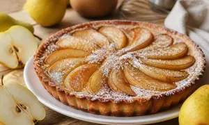 Une délicieuse tarte aux poires, saupoudrée de sucre glace, est présentée sur une assiette blanche. Des poires fraîches sont disposées autour.