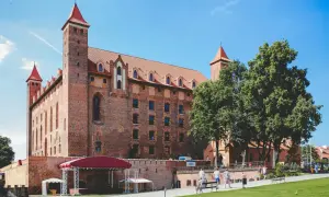 Imponujące ceglane zamki do zwiedzania z czerwonymi dachami i wieżami. Wokół zielony trawnik i drzewa.