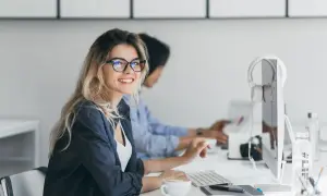 Uśmiechnięta kobieta w okularach pracuje przy komputerze. W tle koleżanka. To obrazek idealnie oddający ducha dyrektywy work life balance.