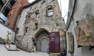 Ein Mann betrachtet die mittelalterliche Steinfassade eines Gebäudes, das Teil des Weltkulturerbes Erfurt ist. Daneben sind alte Steintafeln mit hebräischen Inschriften ausgestellt.