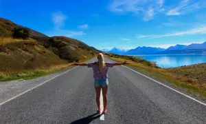 Frau mit ausgebreiteten Armen auf einer Straße, die zu schneebedeckten Bergen und einem blauen See führt. Ein Traum von Work and Travel Weltreise.