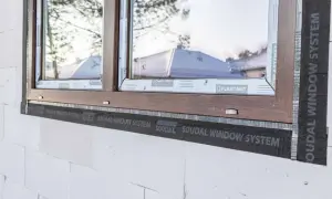 Brązowe okno w trakcie ciepłego montażu z użyciem taśmy SOUDAL WINDOW SYSTEM. Widoczna biała ściana z bloczków.