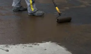 Pracownik w szarym ubraniu z żółtym pasem nakłada wałkiem ciemną masę na betonową powierzchnię. To zbrojenie rozproszone, które wzmacnia konstrukcję.