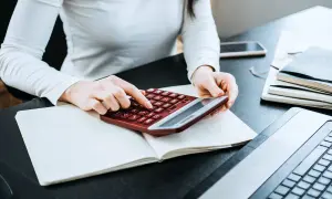 Kobieta liczy wydatki na własne cele mieszkaniowe, przygotowując PIT-39. Na biurku notatnik, kalkulator i laptop.