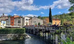 Un pittoresco borghetto mantovano con case colorate, un ponte di legno affollato di gente e un fiume che scorre sotto un cielo azzurro con nuvole bianche.