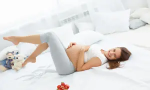 Uśmiechnięta ciężarna kobieta leży na łóżku, głaszcząc brzuch. Obok pluszowe misie i miseczka truskawek. Czasem dokucza jej zespół niespokojnych nóg w ciąży.