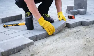 Układanie kostki brukowej na podjazd. Ręce w żółtych rękawiczkach precyzyjnie dopasowują kolejny element. Jaka grubość kostki na podjazd będzie najlepsza?