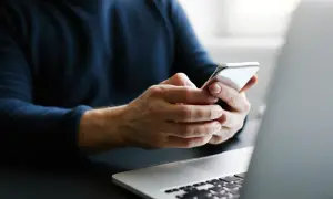 Mężczyzna trzyma telefon, szukając informacji, jak przenieść kontakty na kartę SIM. Obok leży laptop.