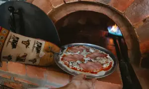 Ręka w rękawicy z krowami wkłada pizzę do pieca szamotowego. Płomień tańczy w głębi pieca.