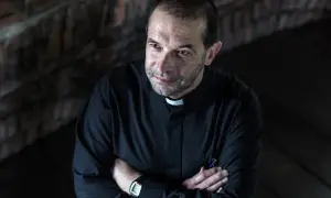 Ksiądz w czarnej sutannie z białym kołnierzykiem, z rękami skrzyżowanymi na piersi. W jego oczach widać troskę, jakby słyszał, że dziewczyna mówi, że jesteś kochany.
