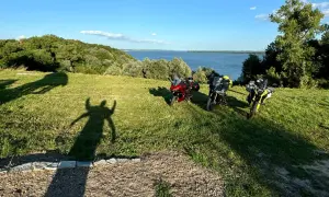 Trzy motocykle na trawiastym wzgórzu z widokiem na jezioro i las.