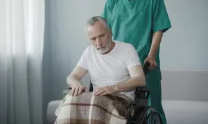 Un uomo anziano in carrozzina, coperto da una coperta, con uno sguardo perso, forse affetto da sindrome locked-in, spinto da un infermiere.
