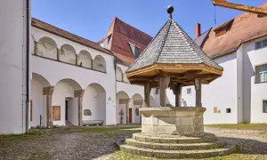 Historischer Brunnen im Innenhof des Oberhaus Passau. Weiße Arkadenbögen und rote Ziegeldächer unter blauem Himmel.