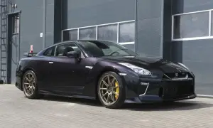Ciemny Nissan R35 z brązowymi felgami i żółtymi zaciskami hamulcowymi stoi przed szarym budynkiem.