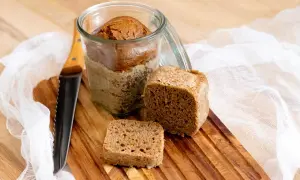 Brot im Glas Rezept: Ein Laib Brot im Glas, daneben ein Stück und ein kleines Quadrat auf einem Holzbrett. Ein Messer liegt daneben.