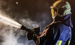 Strażak w hełmie i ubraniu ochronnym trzyma lancę, z której wydobywa się strumień wody.