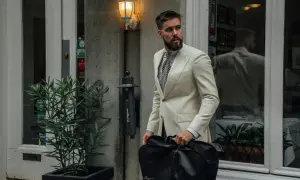 Mann im cremefarbenen Anzug mit gemustertem Hemd und Krawatte, trägt eine schwarze Kleidersacktasche. Ein lässiges Outfit für unterwegs.