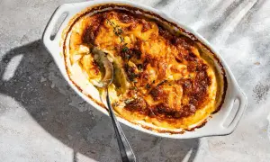 Ein goldbraun gebackenes Kartoffelgratin in einer weißen Auflaufform, garniert mit frischem Thymian. Eine silberne Löffelspitze lugt aus der cremigen Füllung hervor.
