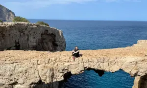 Una mujer sentada en un arco de roca natural, el "cala columpio ibiza", con el mar azul de fondo.